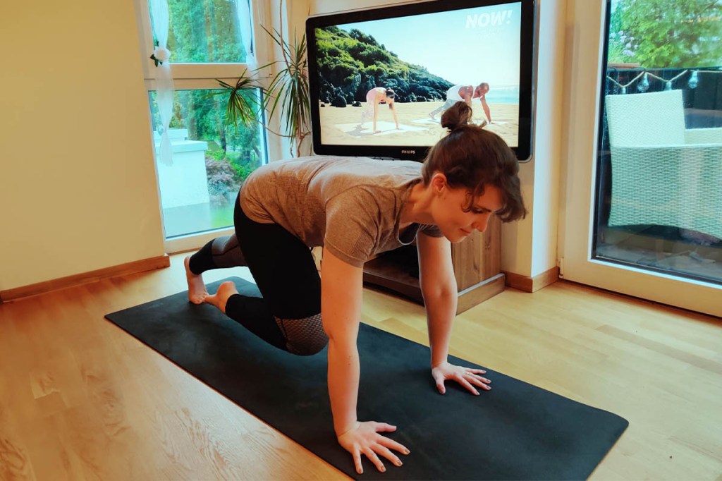Die Testerin macht eine Übung auf einer Yoga-Matte. Dahinter ist ein Fernseher zu sehen, auf dem Kate Hall und Detlef Soost die Übung anleiten.