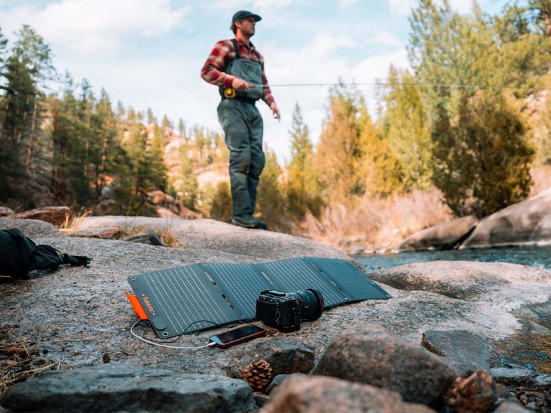 Ein Mann angelt in der Wildnis. Im Vordergrund liegt das Jackery SolarSaga 40 Watt Mini Solarpanel und läd eine Kamera und ein Smartphone auf.