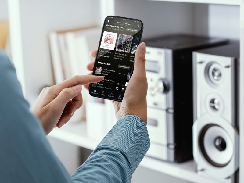 Man hält Smartphone vor Kompakt-Musik-Anlage in beiden Händen. Das Bild soll die Übertragung von Musik per Musikstreamingdienst vom Smartphone an eine kompatible Musikanlage zeigen.
