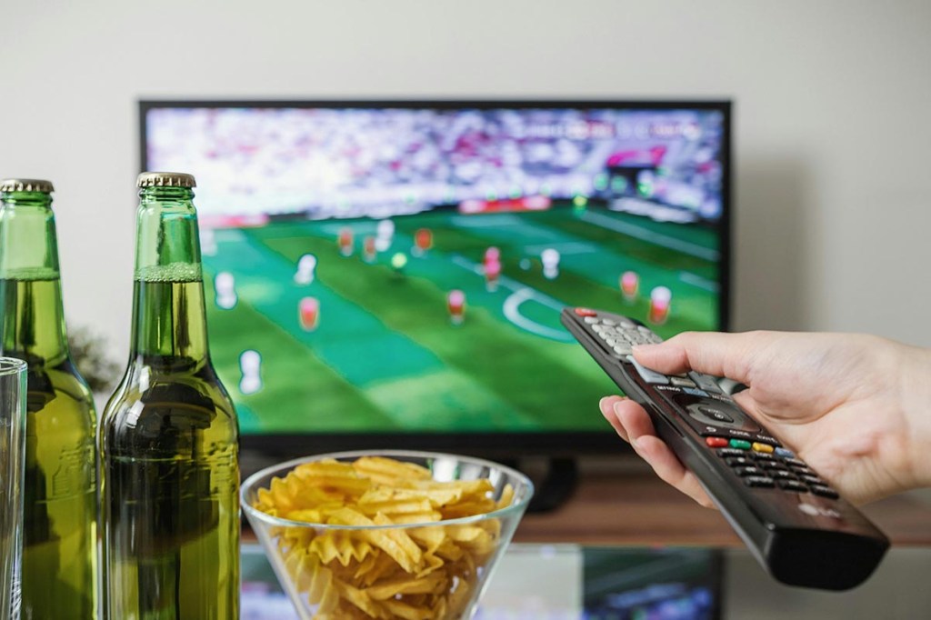 Person mit Fernbedienung in der Hand zeigt auf einen Fernseher.