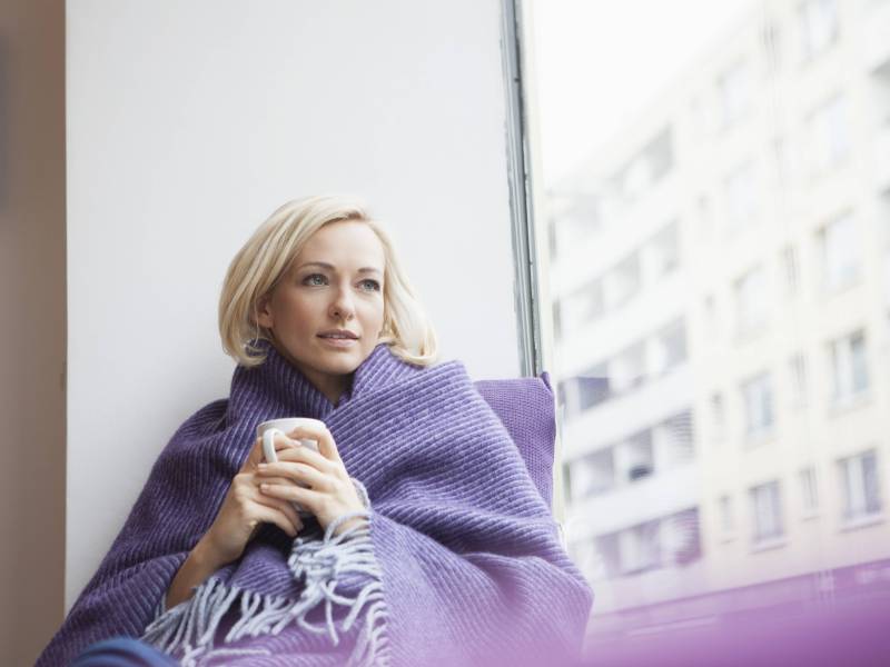 Blonde Frau mit Kurzhaarfrisur sitzt in eine Decke eingehÃ¼llt am Fenster und hÃ¤lt eine Tasse in den HÃ¤nden, wÃ¤hrend sie gedankenversunken aus dem Fenster schaut.