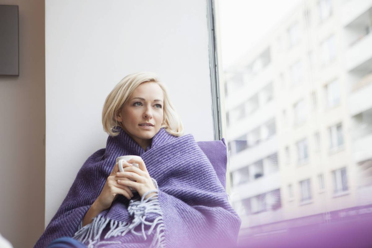 Blonde Frau mit Kurzhaarfrisur sitzt in eine Decke eingehÃ¼llt am Fenster und hÃ¤lt eine Tasse in den HÃ¤nden, wÃ¤hrend sie gedankenversunken aus dem Fenster schaut.