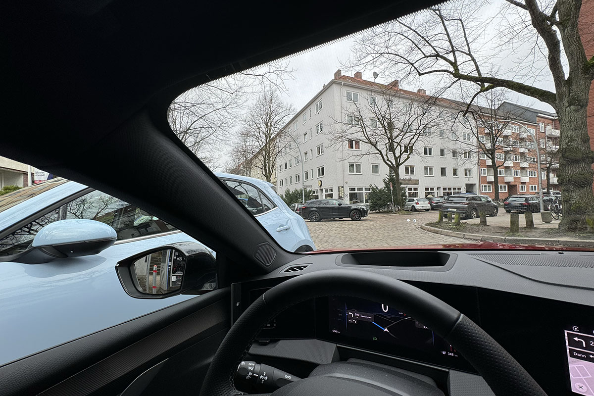 Blick schräg nach links durch die Frontscheibe des E-Autos Opel Astra Electric auf ein Wohnhaus in einer städtischen Umgebung.