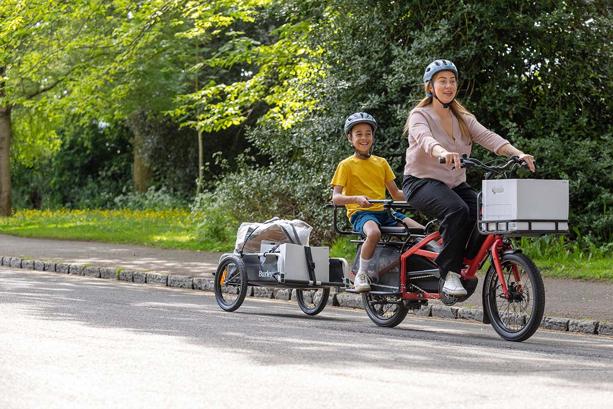 Frau mit einem kompakten Lastenrad, Kind auf Rücksitz und Anhänger