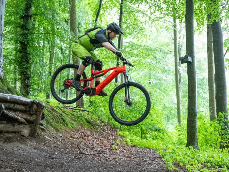 Mann springt mit seinem E-Mountainbike über eine SChanze im Wald