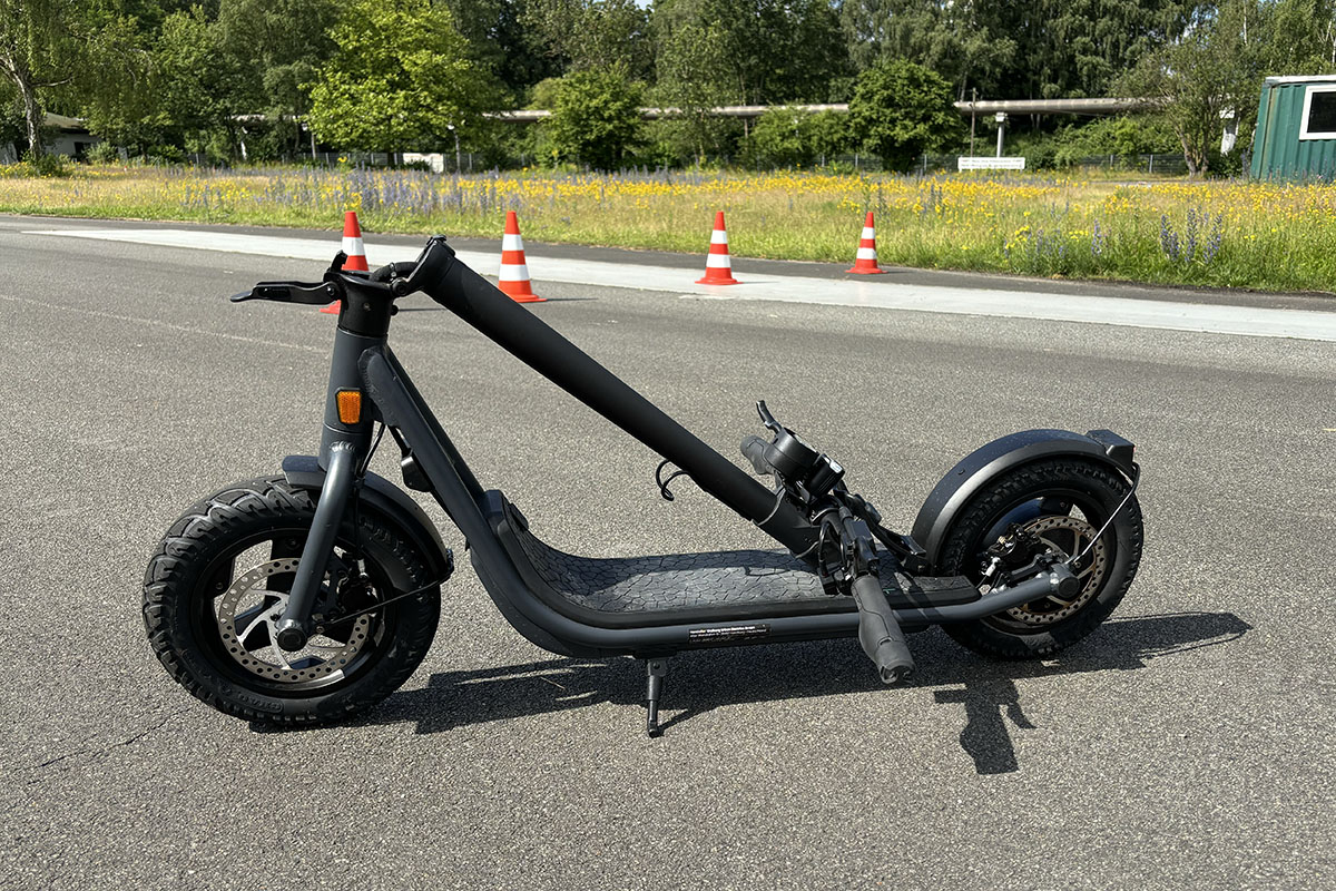 E-Scooter steht zusammengeklappt auf einer Straße vor einer Rasenfläche.