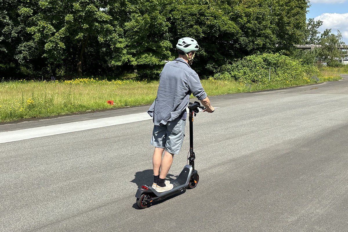 Mann fährt auf einem E-Scooter. Man sieht ihn von hinten an einem Waldstück vorbeifahrend.