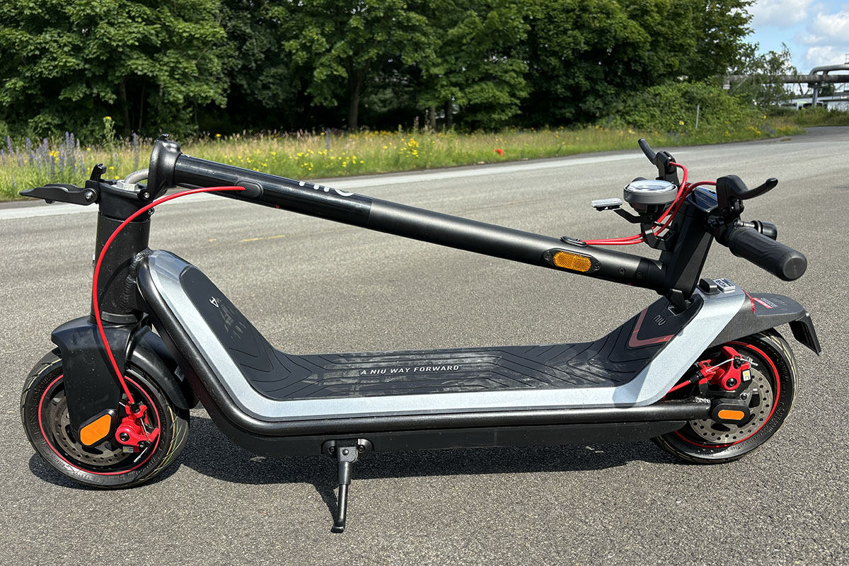 E-Scooter mit eingeklappten Lenker steht im Profil auf einer Straße vor einem Waldstück.