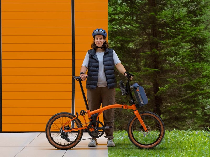 Frau steht hinter ihrem orangenen Faltrad, eine orangene Wand zu einem Teil im HIntergrund