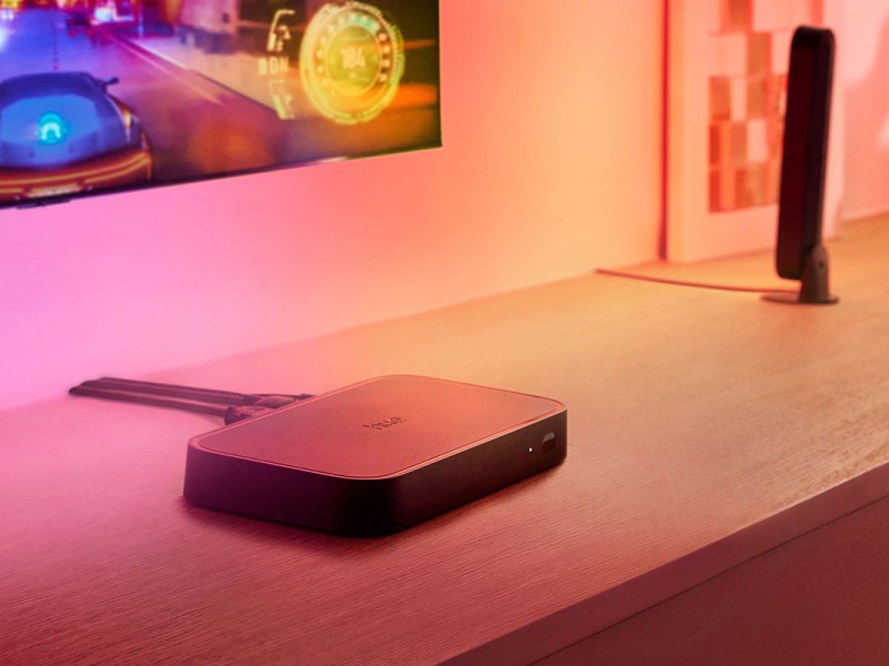 Streaming-Box Philips Hue Play HDMI Sync Box 8K liegt auf einem Sideboard unter einem Flachbild-TV.