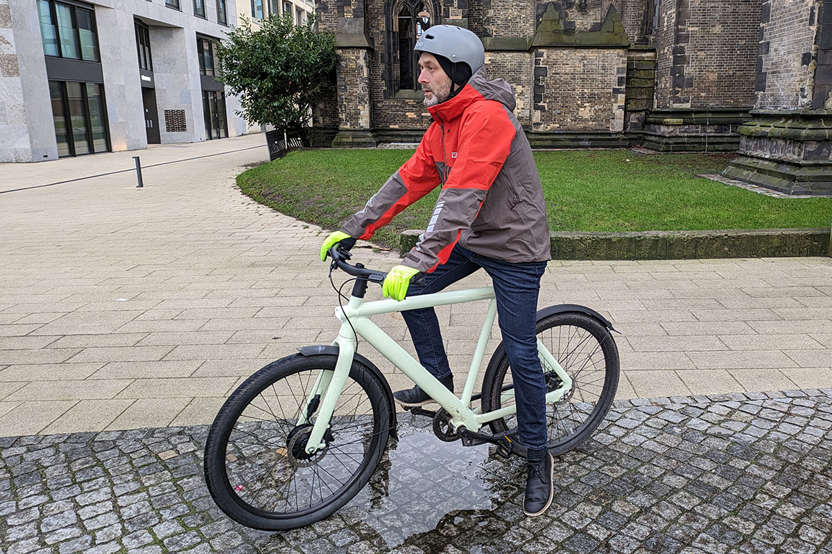 Mann mit grau-roter Jack Wolfskin Morobbia-Regenjacke auf Fahrrad, das in einer Pfütze auf einem gepflasterten städtischen Platz steht.