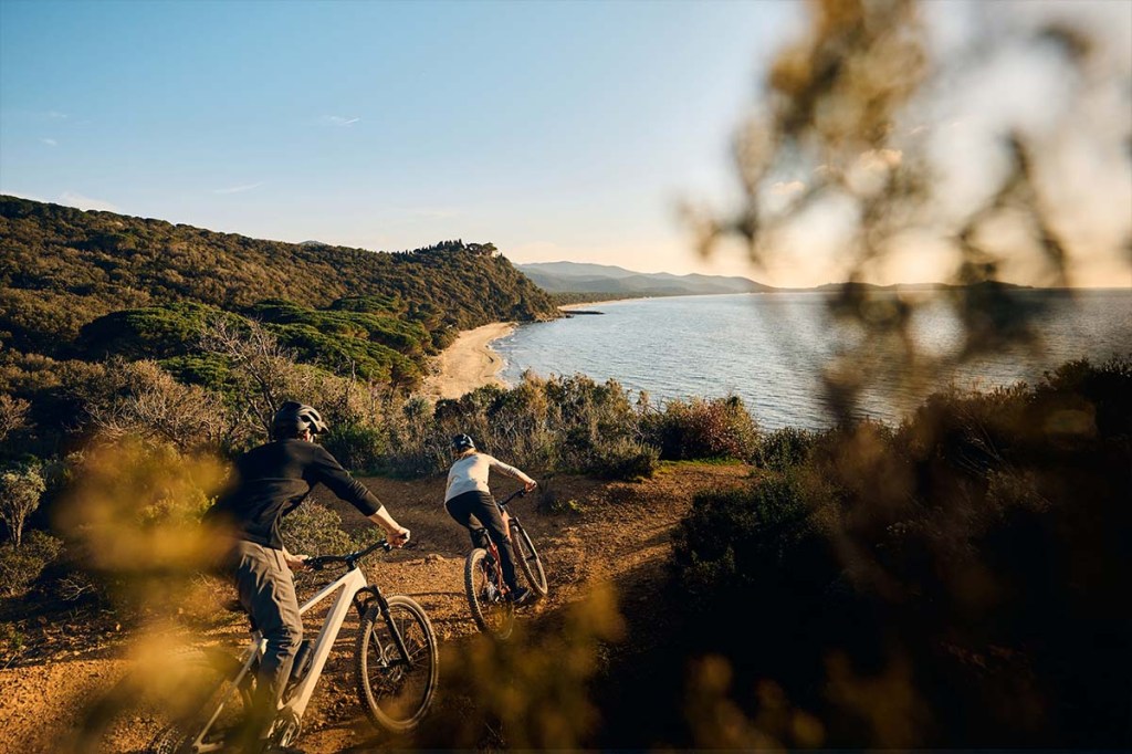 Zwei Personen fahren auf einem Mountainbike durch die Natur, ein See im Hintergrund