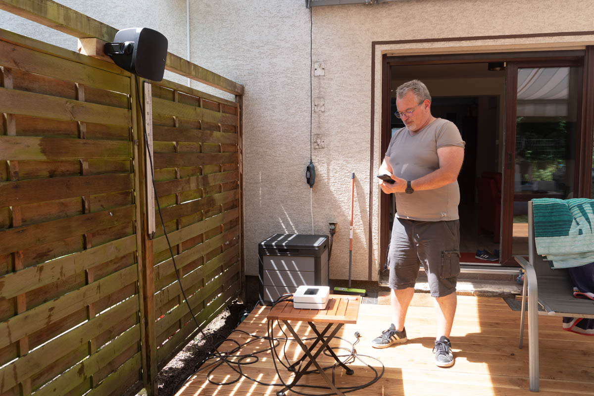 Montage Outdoor-Lautsprecher auf Terrasse an Pergola. Mann steht vor Terrassentür mit Lautsprecher in der Hand.