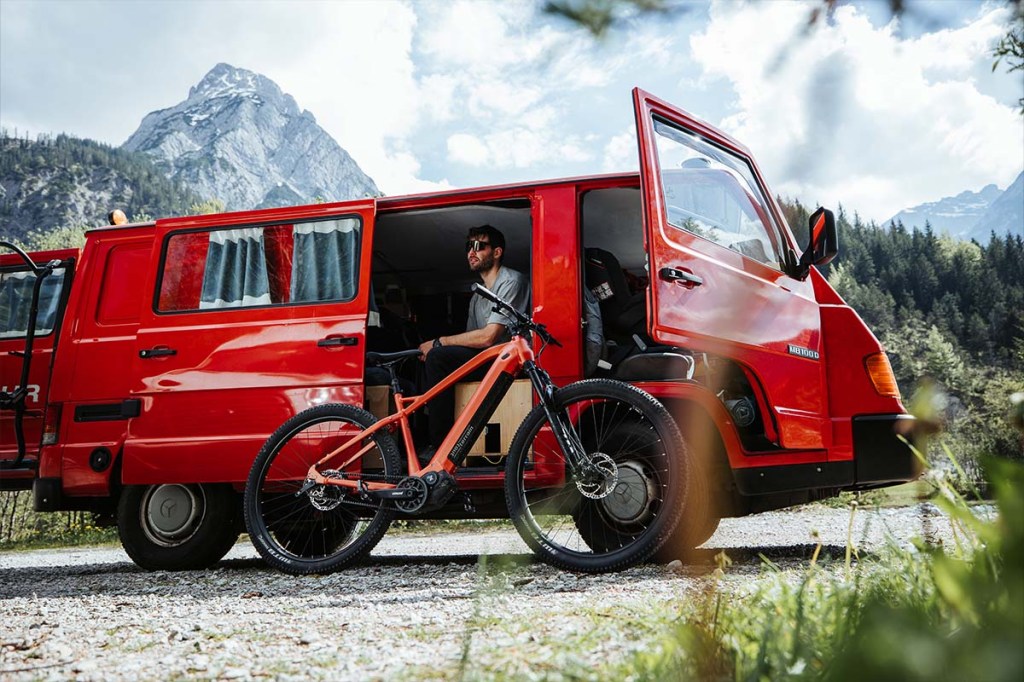 Man sitzt in einem roten Camperbus in der Natur, vor ihm sein E-MTB