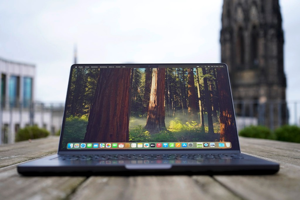 Ein Apple MacBook Steht aufgeklappt mit eingeschaltetem Bildschirm im Freien auf einem Holztisch.