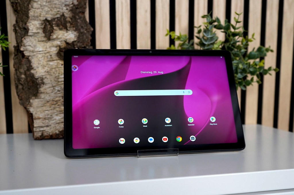 Das T Tablet auf einer weißen Ablage mit Pflanzen im Hintergrund vor einer Holzwand. Der Bildschirm-Hintergrund ist pink-schwarz.