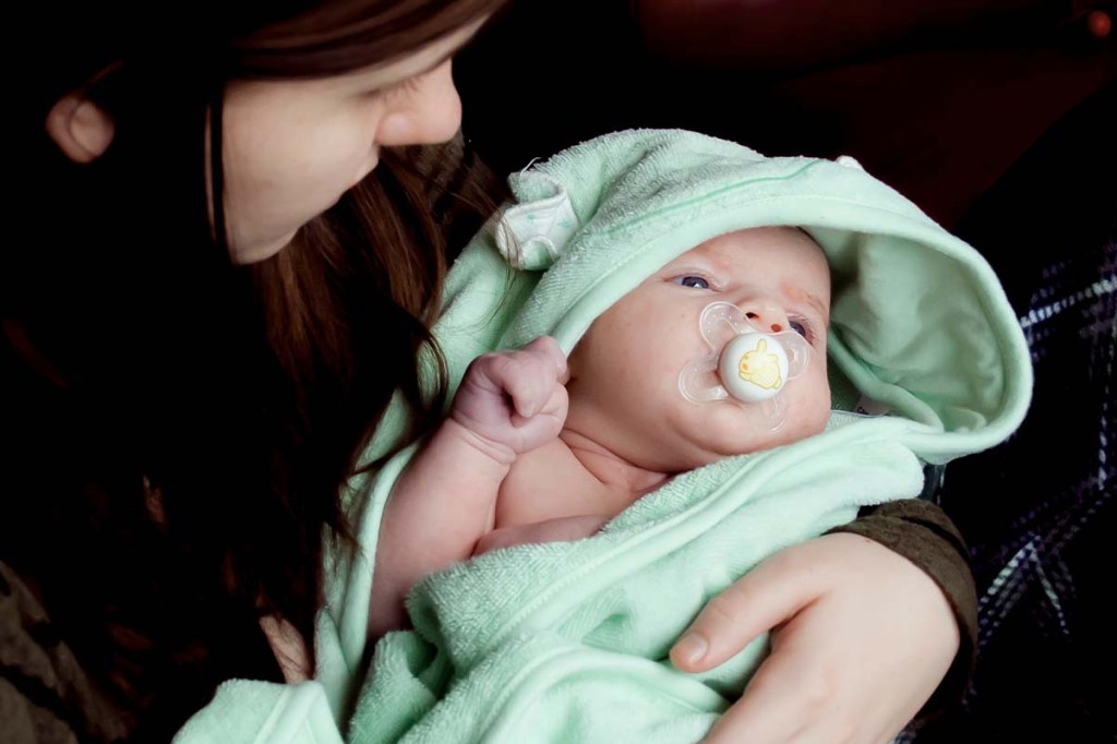 Baby mit Schnuller im Mund und umhüllt in einem grünen Badetuch auf dem Arm einer Frau