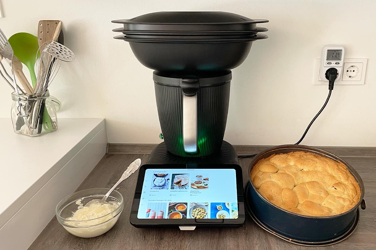 Der Thermomix TM7 mit Varoma-Aufsatz und eingeschaltetem Display in Küchenumgebung. Daneben stehen ein Kuchen und Schlagsahne. Der Stecker steckt zudem in einem Leistungsmessgerät.