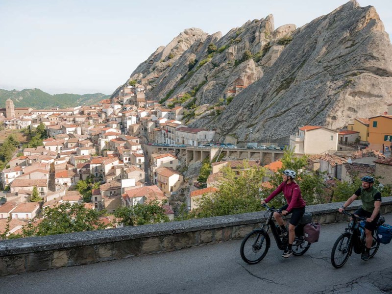 zwei Radfahrer fahren eine Straße entlang, ein Dorf in einem Tal sowie Berge im Hintergrund