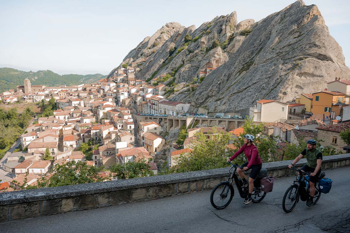 zwei Radfahrer fahren eine Straße entlang, ein Dorf in einem Tal sowie Berge im Hintergrund