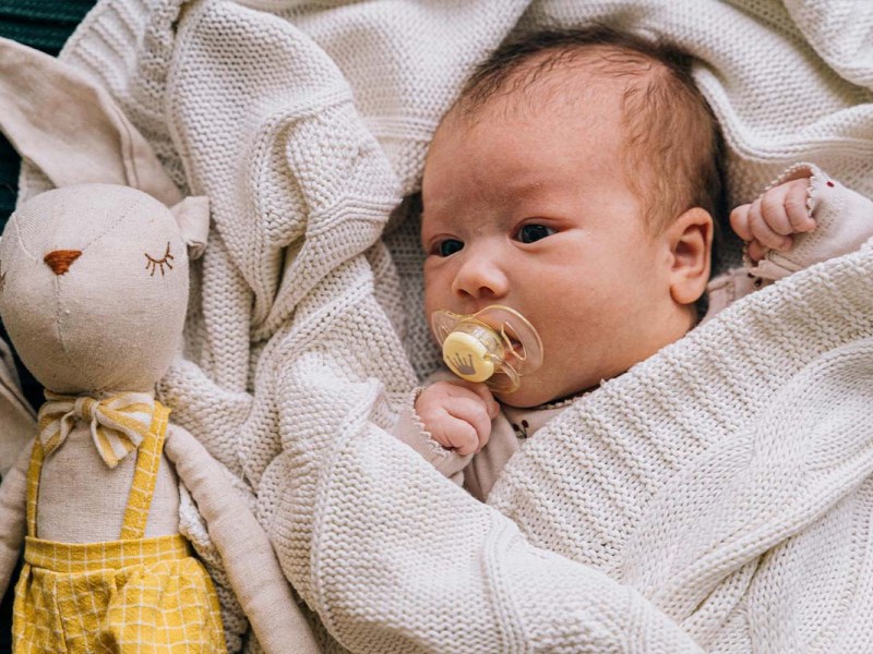 Baby mit Schnuller im Mund in einer Decke eingehüllt, neben ihm liegt ein Kuschelhase