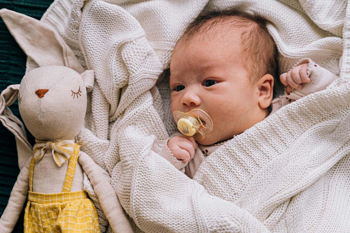 Baby mit Schnuller im Mund in einer Decke eingehüllt, neben ihm liegt ein Kuschelhase