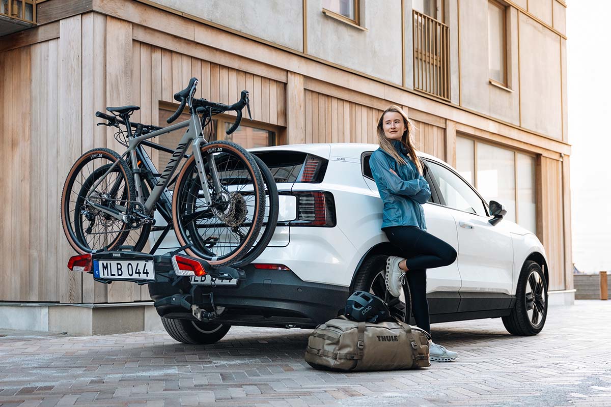 Frau steht vor einem weißen Auto, an diesem ist ein Heckträger mit zwei Gravelbikes befestigt
