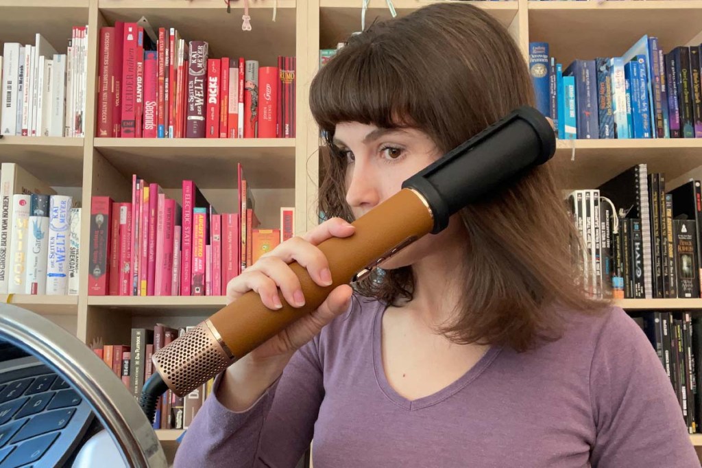Eine Frau sitzt vor einem bunten Bücherregal und glättet sich mit dem Dreame Airstyle Pro und dem Anti-Frizz-Aufsatz die Haare.