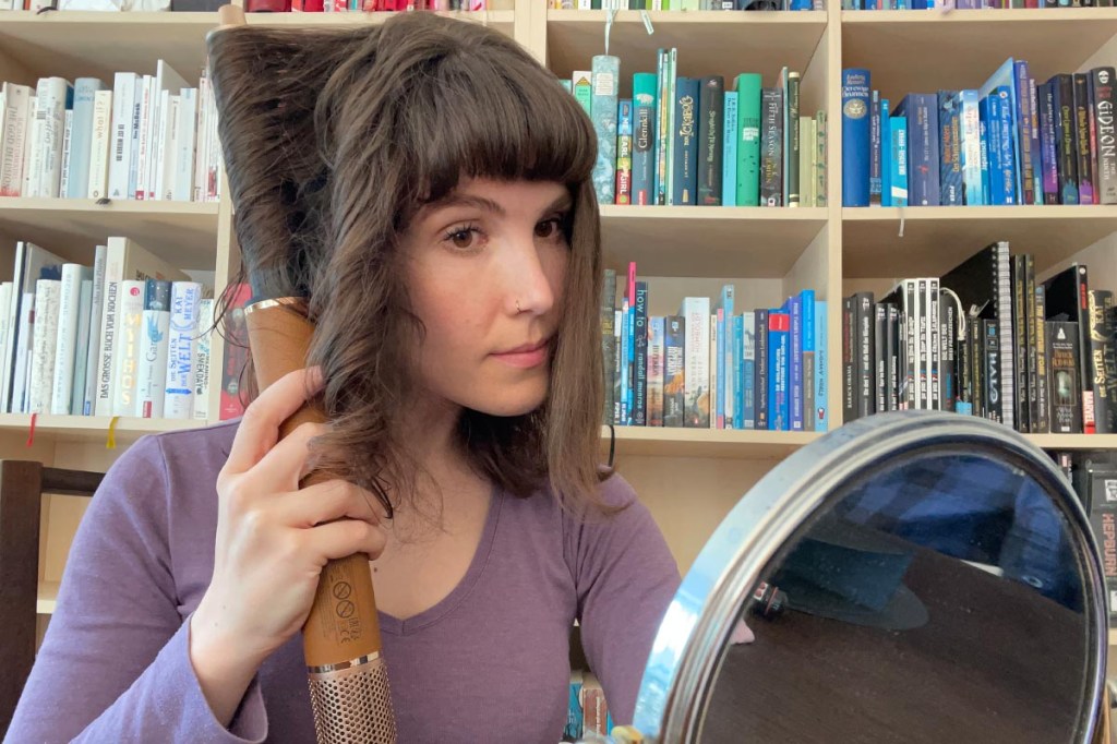 Eine Frau sitzt vor einem bunten Bücherregal und lockt sich mit dem Dreame Airstyle Pro und dem Lockenstab-Aufsatz die Haare.