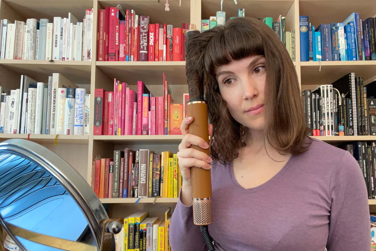 Eine Frau sitzt vor einem bunten Bücherregal und lockt sich mit dem Dreame Airstyle Pro und dem Lockenstab-Aufsatz die Haare.