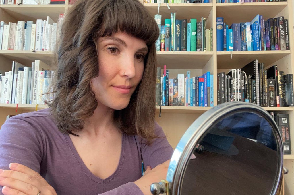 Eine Frau sitzt vor einem bunten Bücherregal und betrachtet ihre Frisur in einem Kosmetik-Spiegel. Eine Seite ihrer Haare ist glatt, eine lockig.