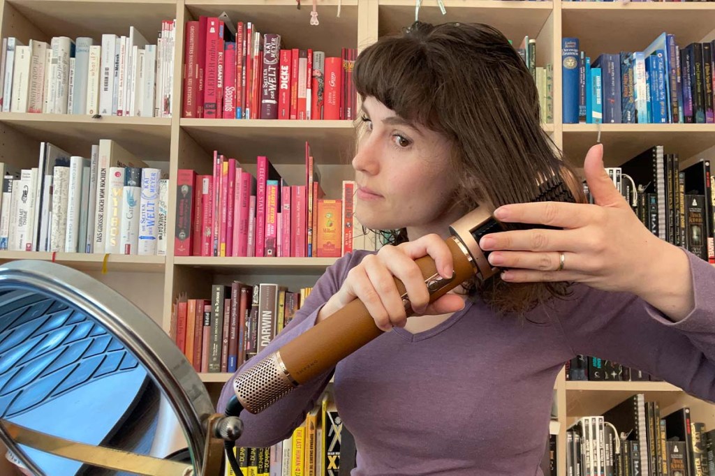 Eine Frau sitzt vor einem bunten Bücherregal und trocknet sich mit dem Dreame Airstyle Pro und dem Bürsten-Aufsatz die Haare.