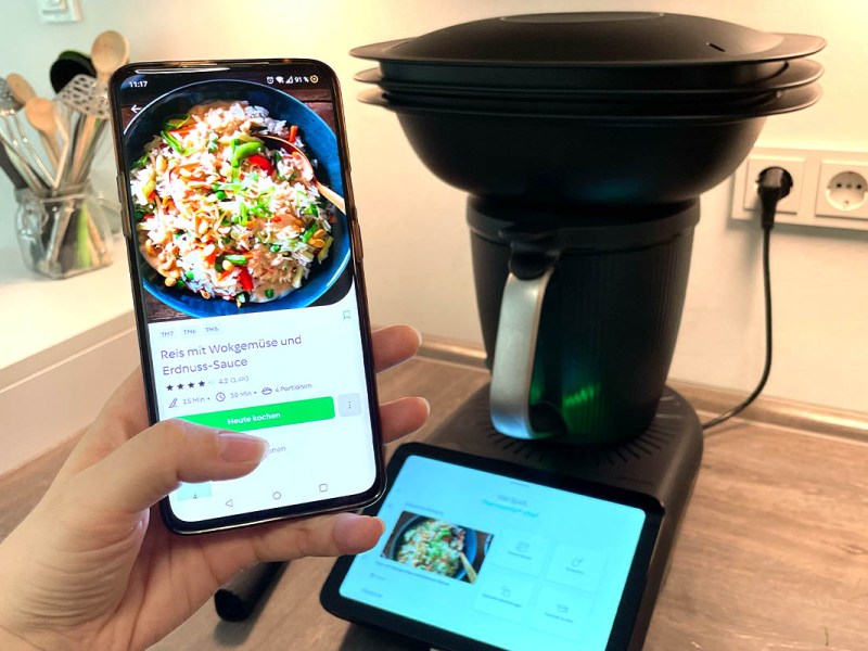 Eine Hand hält ein Smartphone mit Cookidoo-App in die Kamera. Dahinter der Thermomix TM7 auf einer Küchenarbeitsplatte.